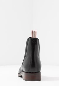 Bottine en cuir noir avec talon en bois, panneaux élastiques latéraux et une languette de tirage rayée rouge et crème portant l'inscription "GANT".