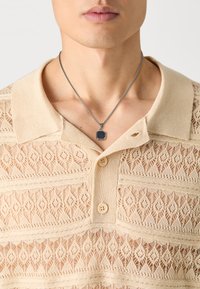 Beige strikket polo-skjorte med blonder-lignende mønster og tre knapper. En sølvkjede med firkantet anheng henger over kragen.