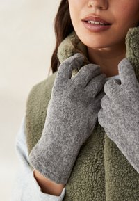 Guanti di lana grigia con una finitura morbida e testurizzata, indossati mentre si sistema un gilet in pile verde oliva. Attenzione al comfort e al calore.