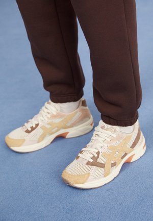 Persoon die witte en beige mesh sneakers draagt met bruine joggingbroek, staand op een blauwe ondergrond.