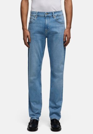 Mand iført lyseblå jeans med lige ben, hvid skjorte stoppet ned i bukserne og sorte loafers, står med hænderne afslappet ned langs siderne.