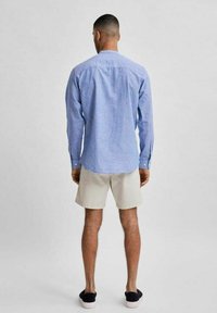 Camicia a maniche lunghe a quadretti blu, tessuto leggero, colletto a polo, abbinata a pantaloni corti beige e scarpe scivolate scure, vista da dietro.