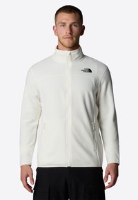 Chaqueta de forro polar blanca con cuello alto, cremallera frontal y dos bolsillos laterales. Presenta un logo negro en el lado izquierdo del pecho. Textura suave.