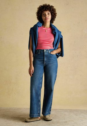 Junge Frau mit lockigem Haar, die ein rot-weiß gestreiftes Shirt, blaue Jeans, eine Jeansjacke über den Schultern und bequeme beige Schuhe trägt.