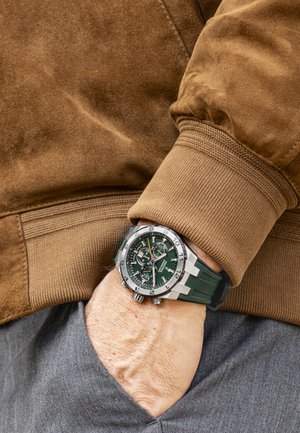 Montre à cadran vert avec un boîtier en acier inoxydable argenté, fonctionnalités chronographe et bracelet en caoutchouc vert, portée sur un poignet partiellement visible.
