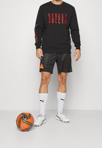 Sort sweatshirt med rød teksten "STREET SOCCER", sorte shorts med orange accenter, hvide sokker og fodboldstøvler. Orange og sort fodbold på jorden.