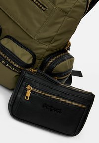 Zaino trapuntato verde oliva con numerose tasche nere con zip e dettagli in oro, accompagnato da una pochette in pelle nera texturizzata con logo.