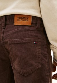 Brown corduroy pants featuring a leather Tommy Hilfiger label and a small logo detail in red, white, and blue on the pocket.