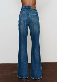 Tmavomodré rozšírené džínsy s vysokým pásom, päť vreciek a koženou nášivkou na zadnej strane. Oblečený lem pridáva textúru.