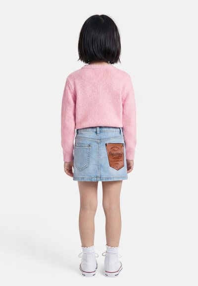 Παιδί με κοντά μαύρα μαλλιά που φοράει ροζ πουλόβερ, φούστα από ανοιχτό μπλε τζιν με μεγάλο δερμάτινο σημείο στην πίσω τσέπη, λευκές κάλτσες και αθλητικά παπούτσια.