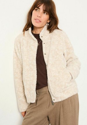 Beige, plush fleece jacket with a high collar, snap closures, and side pockets, paired with a dark top and brown textured pants.