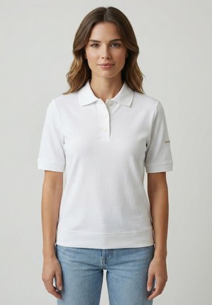 Donna vista frontalmente che indossa una polo bianca a maniche corte e jeans azzurri chiari su uno sfondo grigio chiaro uniforme.