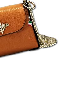 Sac en cuir brun avec une bandoulière en chaîne dorée, présentant un accent abeille sur le rabat et un petit charme drapeau italien. Texture lisse.