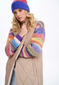 Beżowa pikowana kamizelka z bocznymi kieszeniami, w zestawie z wielokolorowym dzianinowym swetrem w paski w różnych kolorach oraz fioletową dzianinową czapką.
