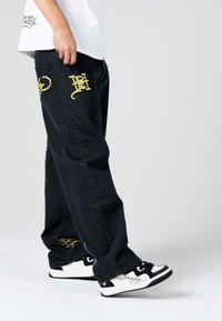 Los pantalones de mezclilla oscura presentan diseños bordados en amarillo en los bolsillos traseros. Se combinan con zapatillas de deporte en blanco y negro, mostrando un contraste de color.