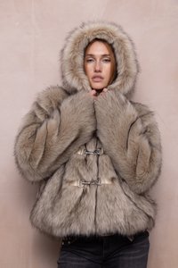 Woman wearing a thick, fur hooded coat with metal clasps, holding the coat close around her face, standing against a plain beige background.