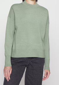 Hellgrüner Pullover aus weicher Strickware mit hohem Kragen, gerippten Bündchen und lockerer Passform. Kombiniert mit dunkelgrauen Jeans.