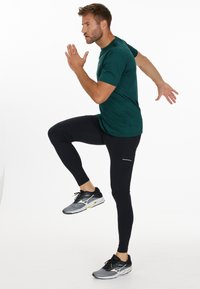 Groene sport t-shirt gecombineerd met zwarte legging. De legging heeft een reflecterende strook. Zwarte en grijze hardloopschoenen met witte accenten.