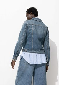 Veste en jean à coupe ajustée, teinte bleu moyen, col classique et détails à boutons. Associée à une chemise plissée rayée en dessous.
