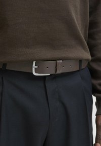 Brown leather belt with a silver-colored buckle and a textured finish, worn over navy trousers and a dark brown sweater.