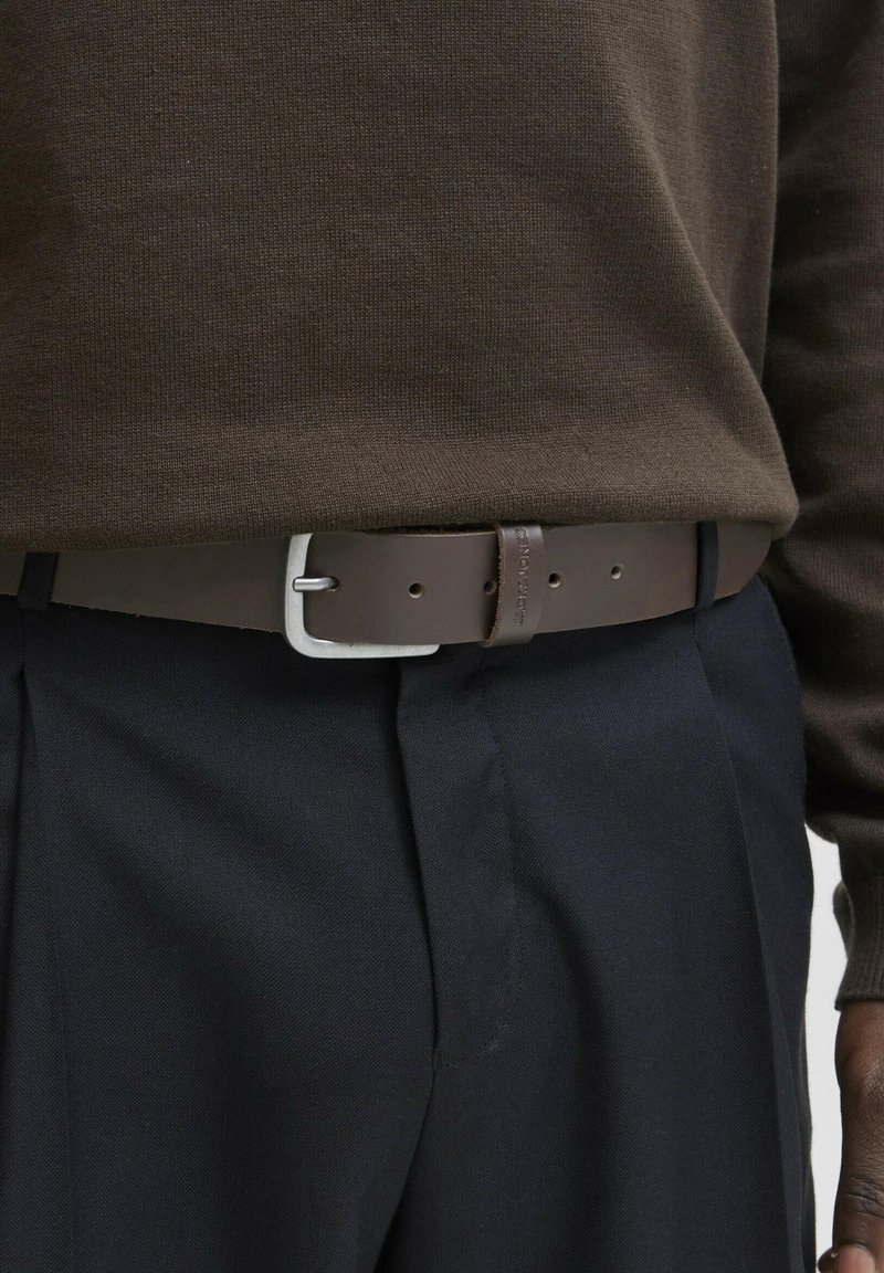 Brown leather belt with a silver-colored buckle and a textured finish, worn over navy trousers and a dark brown sweater.