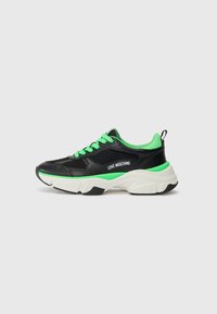 Zwarte sneaker met groene accenten, meshy en leren materialen. Dikke witte zool, logo op de zijkant en contrasterende veters.