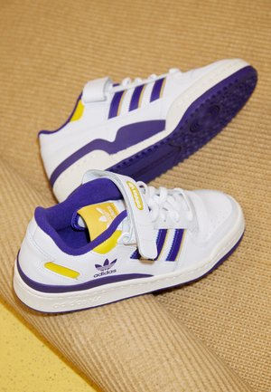 Białe trampki Adidas z fioletowymi i żółtymi akcentami, z trzema paskami, rzepem i logo, umieszczone na fakturowanej beżowej powierzchni.