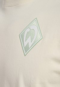 Cremefarvet T-shirt med en struktureret mintgrøn lap og et hvidt logo. Stoffet ser blødt og glat ud uden synlige mønstre.