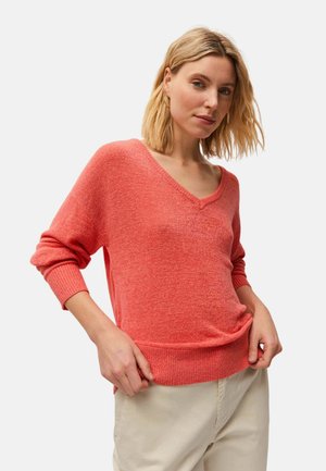 Mujer con cabello rubio, vestida con suéter coral de cuello en V y pantalones beige, de pie con las manos cerca de la cintura contra un fondo liso.