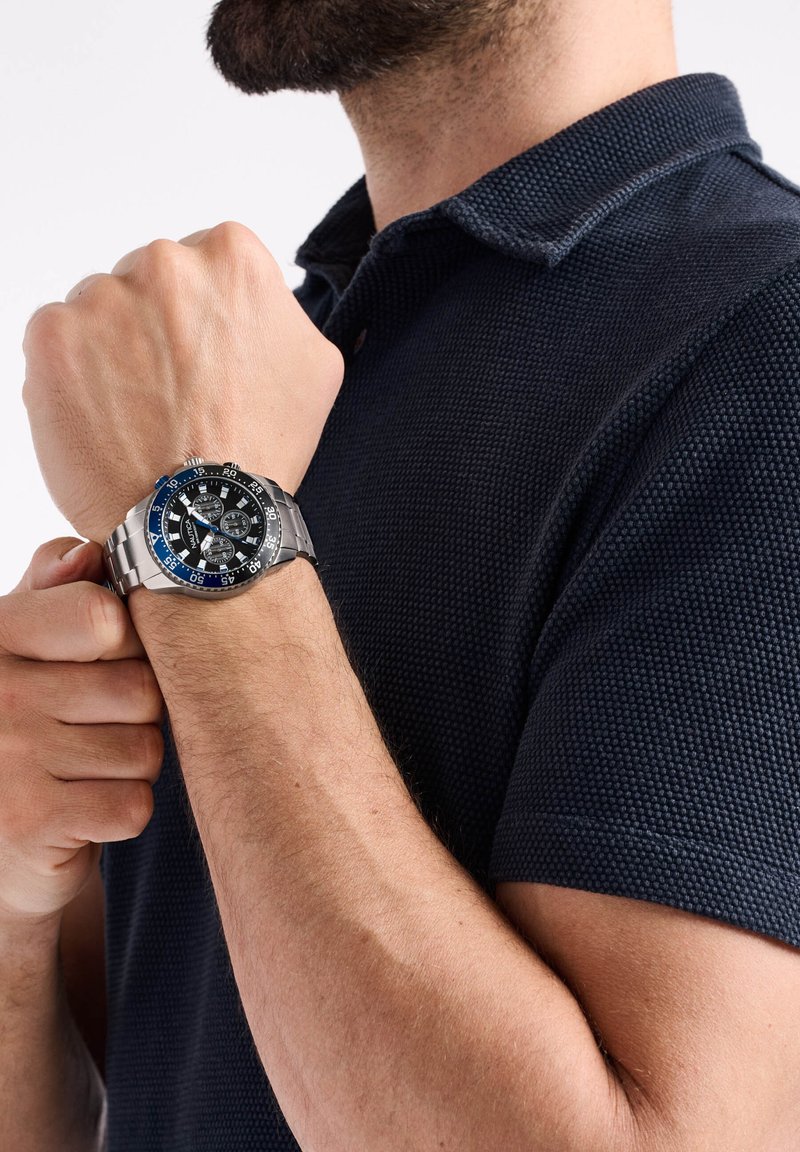 Reloj de acero inoxidable plateado con esfera negra y acentos azules, que presenta múltiples subesferas y un bisel taquimétrico. Lleva puesto en una muñeca.