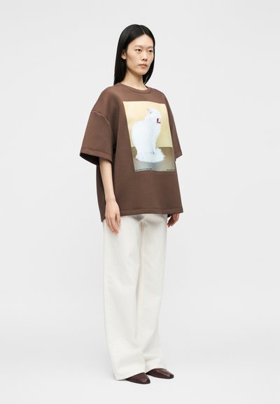 Γυναίκα με μακριά μαύρα μαλλιά που φοράει μεγάλη καφέ φούτερ με λευκό σχέδιο γάτας, λευκό φαρδύ παντελόνι και καφέ μυτερά παπούτσια, όρθια.