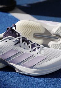Paire de chaussures de sport blanche et violette avec tige en mesh et motif rayé sur une surface texturée bleue.