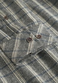 Cămașă în carouri gri și alb cu model de herringbone, având nasturi maro, textură din bumbac și detaliu cu manșetă pliată.