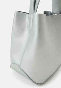 Róg miękkiej szarej torby typu tote z widocznym przeszyciem i gładką fakturą na białym tle.