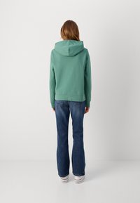 Felpa verde con cappuccio, realizzata in morbido misto cotone. Abbinata a jeans a zampa d'elefante blu scuro. Design semplice, senza loghi o accenti visibili.