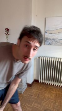 Jeune homme avec moustache penché en avant à l'intérieur, sol en parquet, radiateur blanc, œuvre d'art murale abstraite et fleurs rouges dans un vase en arrière-plan.