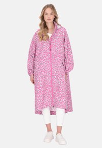 Dlouhý kabát s růžovým leopardím vzorem, zapínáním na zip, měkkou texturou a elastickými manžetami, nošený přes bílé tričko a bílé legíny.