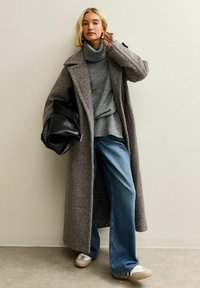 Un abrigo largo de lana gris sobre un suéter de cuello alto gris, combinado con jeans de pierna ancha azul y zapatillas blancas. Un bolso negro oversized completa el look.