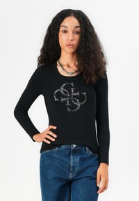 Svart langermet topp med en teksturert logo i sølvdetaljer. Kombinert med høyt liv blå denimbukser.