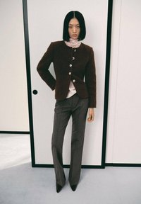 Veste texturée marron avec des boutons en argent, superposée à un top rose. Un pantalon gris ajusté et des talons noirs pointus complètent la tenue.
