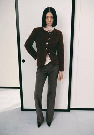 Giacca marrone con texture e bottoni argentati, indossata sopra una maglietta rosa. I pantaloni grigi su misura e i tacchi neri a punta completano l'outfit.