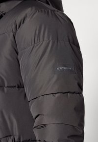 Veste matelassée noire présentant des sections matelassées, une texture lisse et un patch logo bien visible sur la manche.