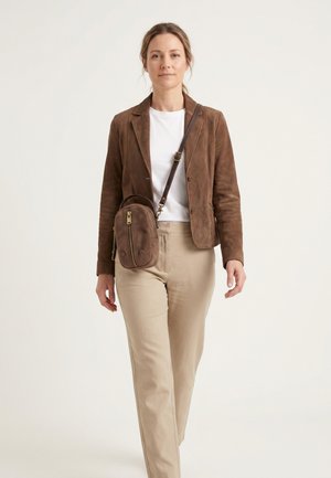 Vrouw loopt naar voren, draagt een bruine suède blazer, wit overhemd, beige broek en een kleine bruine crossbodytas met ritssluiting details.