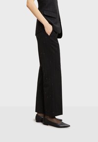 Černé pruhované kalhoty se střihem rovného nohavice, vyrobené z texturované látky. V páru s černými balerínami s špičatou špičkou, nošeno s rukama v kapsách.