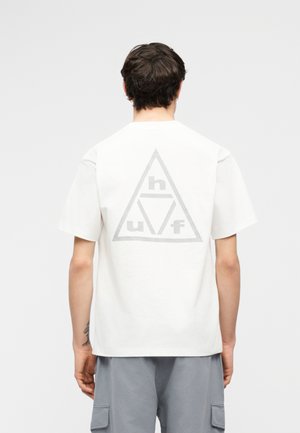 Άνδρας με κοντά σκουρόχρωμα μαλλιά φοράει φαρδιά λευκή μπλούζα με γκρι τριγωνικό λογότυπο και τα γράμματα "h", "u", "f" στην πλάτη, καθώς και γκρι σορτς cargo.