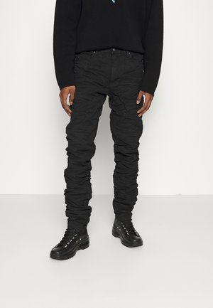 Personne portant un pantalon noir froissé, des bottes montantes noires à lacets et un pull noir à manches longues, debout sur un fond blanc.