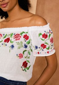 White off-the-shoulder top with floral embroidery in reds, blues, and greens. Features textured fabric and a ruffled hem.