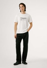Baltas T-krekls ar melnu grafisko druku, kurā redzams "PEPE JEANS LONDON." Saskaņots ar melnajiem taisnajiem biksēm un melnajiem apaviem.