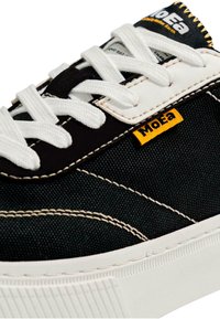 Fekete vászon sneaker fehér fűzővel, sárga logó részlettel és fehér gumitalppal. Kontrasztos varrás és texturált anyag jellemzi.