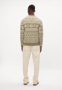 Maglione lavorato a maglia in verde oliva con motivi geometrici bianchi, colletto rotondo, polsini e orlo a costine. Abbinato a pantaloni chiari.